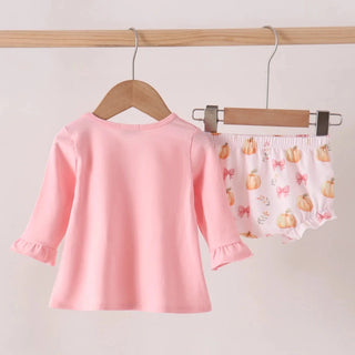 A two-piece baby clothing set hanging on wooden hangers from a wooden rod. The set includes a light pink long-sleeve top with ruffled cuffs and matching light pink bloomers with a pumpkin and bow pattern.