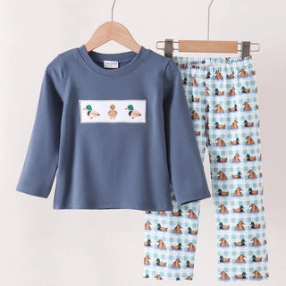 A two-piece children's pants set hanging on wooden hangers against a plain light background. The set includes a slate blue long-sleeve top with a white rectangular chest patch showing three cartoon ducks, and matching light blue pants covered in a repeating duck print.