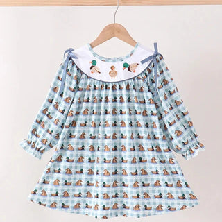 A light blue and white gingham long-sleeve baby dress with a repeating dog print, featuring a white Peter Pan collar decorated with embroidered ducks, hanging on a wooden hanger.