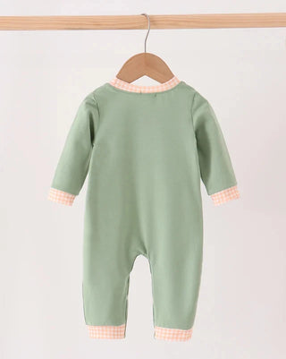 A sage green baby jumpsuit hanging on a wooden hanger from a light wood bar, featuring peach and white gingham trim at the collar, cuffs, and hem.