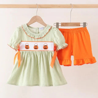A toddler girl's two-piece Halloween outfit hanging on wooden hangers against a light beige wall. The set includes a light sage green short-sleeve dress with orange ruffle trim, a white chest band with three embroidered orange jack-o'-lantern faces, and matching bright orange shorts with a ruffled hem.