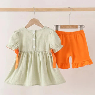 A toddler girl's two-piece outfit set hanging on wooden hangers against a plain light pink wall. The set includes a sage green short-sleeve dress with white buttons and orange side bows, paired with bright orange ruffle-hem shorts.