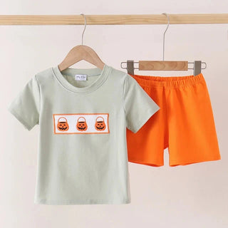 A children's two-piece outfit set hanging on wooden hangers against a plain light background. The set includes a light sage green short-sleeve T-shirt with a rectangular chest graphic showing three orange jack-o'-lantern faces in buckets, and bright orange shorts.