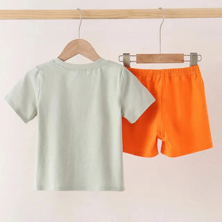 A children's two-piece outfit hanging on wooden hangers from a wooden rod. The set includes a short-sleeve crewneck t-shirt in a pale sage green and matching shorts in a bright orange.