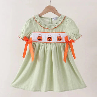 A light sage green dress with orange trim, featuring a white chest band decorated with three orange jack-o'-lanterns and orange ribbon bows on the sleeves, hanging on a wooden hanger against a plain beige wall.