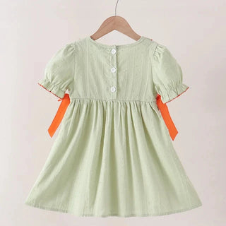 A light sage green dress with short puff sleeves, white buttons, and orange ribbon accents on the sides, hanging on a wooden hanger against a plain beige background.