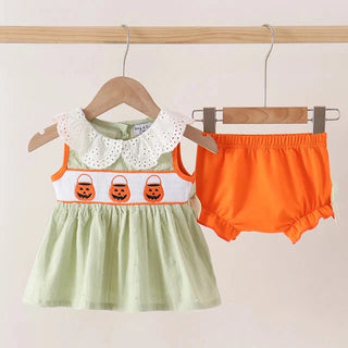 A baby girl's two-piece Halloween outfit hanging on wooden hangers against a light beige wall. The set includes a light sage green smocked dress with a white eyelet Peter Pan collar and an orange chest band decorated with three embroidered jack-o'-lantern buckets, paired with bright orange ruffle-bloomer shorts.