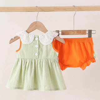 A baby girl's two-piece outfit set hanging on wooden hangers from a wooden rod. The set includes a light sage green sleeveless dress with a white eyelet Peter Pan collar and white buttons, and a pair of bright orange bloomers.
