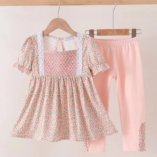 A toddler girl's two-piece clothing set hanging on wooden hangers against a plain beige wall. The set includes a short-sleeve dress with a pink, green, and white floral print, white lace trim at the neckline, and matching pink leggings with a floral patch detail on the lower leg.