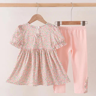 A toddler girl's two-piece outfit hanging on wooden hangers against a plain light background. The set includes a short-sleeve dress with a small multicolor floral print and matching light pink leggings.