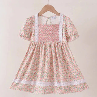 A short-sleeve girl's dress with a pink, green, and white floral print, featuring a smocked bodice, white lace trim, and a gathered skirt, hanging on a wooden hanger against a light beige wall.