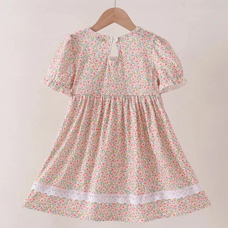 A short-sleeve girl's dress with a pink, green, and white floral print, hanging on a wooden hanger against a plain beige wall.