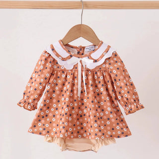 A toddler girl's long-sleeve romper dress in terracotta orange with a white and black floral pattern, featuring a white ruffled Peter Pan collar and bow, hanging on a wooden hanger.