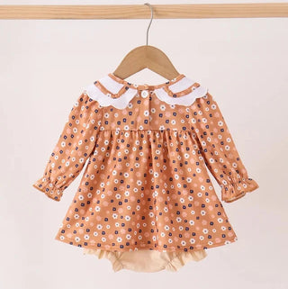 A toddler girl's long-sleeve dress in a warm peach-orange color, decorated with a white and navy blue floral pattern. It features a white ruffled Peter Pan collar and a beige ruffled hem, hanging on a wooden hanger against a solid light beige background.