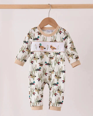 A baby long-sleeve romper hanging on a wooden hanger, featuring an all-over duck and reeds print on an off-white background, with a central horizontal panel showing two ducks.