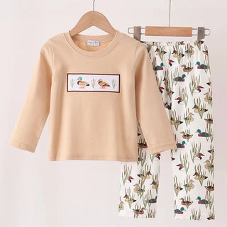A children's two-piece outfit set hanging on wooden hangers against a plain light background. The set includes a long-sleeve beige top with a rectangular duck-themed chest graphic and matching white pants covered in a colorful duck, cactus, and bird print.