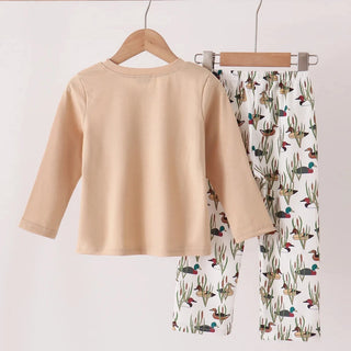 A two-piece children's outfit hanging on wooden hangers from a wooden rod. The set includes a light beige long-sleeve top and white pants with a colorful toucan and cactus print.