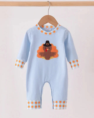 A baby long-sleeve light blue romper hanging on a wooden hanger, featuring an applique turkey design on the chest and orange-and-white checkered trim at the collar, cuffs, and leg openings.