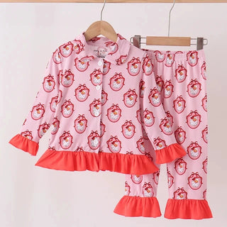 A two-piece children's pajama set hanging on wooden hangers against a plain light background. The set includes a long-sleeve button-front top and matching pants, both in light pink with a repeating red santa print and bright coral-red ruffled trim at the hem and cuffs.