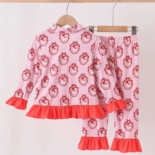 A two-piece children's pajama set hanging on wooden hangers against a plain light background. The set includes a long-sleeve top and matching pants in light pink with a repeating red and white santa pattern, finished with bright coral-red ruffled trim at the cuffs and hem.