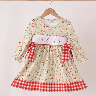 A long-sleeve dress with a cream base, red and green chili pepper print, a white smocked chest panel with embroidered details, and red gingham trim on the waist and hem, hanging on a wooden hanger against a plain white background.