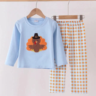 A two-piece children's pants set hanging on a wooden hanger bar. The set includes a light blue long-sleeve top with a turkey graphic and matching orange-and-white gingham check pants.