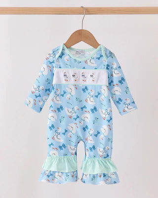 A baby long-sleeve romper in light blue with a white chest panel, featuring a playful print of geese, bows, and stars. The romper has ruffled trim at the cuffs and is displayed on a wooden hanger against a plain off-white background.