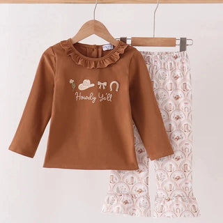 A toddler girl's two-piece outfit hanging on wooden hangers against a light beige wall. The set includes a brown long-sleeve top with a ruffled neckline and western-themed embroidery reading 'Handy Yell' with a cowboy hat, bow tie, and horseshoe, and matching light pink patterned pants with a ruffled hem.