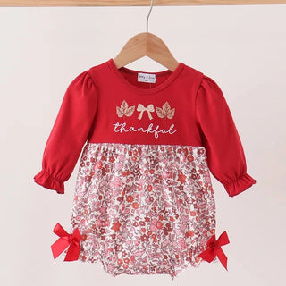 A toddler girl's long-sleeve romper dress hanging on a wooden hanger against a light beige background. The top half is solid red with 'thankful' and leaf graphics, and the bottom half is a red and white floral patterned bloomers section with red ribbon bows on the sides.