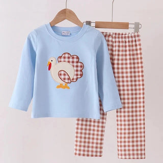 A two-piece children's pants set hanging on wooden hangers against a plain light background. The set includes a light blue long-sleeve top with a turkey applique on the front and matching red-and-white gingham pants.