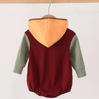 A baby hooded romper hanging on a wooden hanger from a wooden clothes rack. The romper features a burgundy body, sage green sleeves, and a contrasting orange hood.