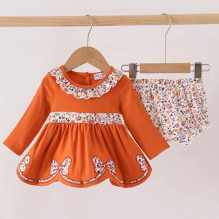 A baby girl's two-piece outfit set consisting of a long-sleeve orange dress with a white floral ruffled collar, a matching floral waistband, and butterfly embroidery on the skirt, paired with matching floral bloomers, hanging on wooden hangers against a plain light background.