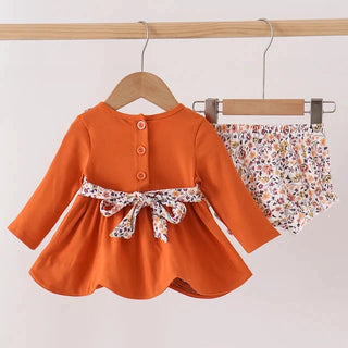 A toddler girl's two-piece outfit hanging on wooden hangers against a plain white wall. The set includes a long-sleeve orange dress with a floral-patterned waist tie and matching floral bloomers.