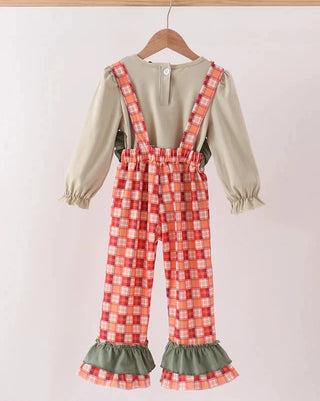 A toddler girl's two-piece outfit hanging on a wooden hanger against a light pink background. The set includes a beige long-sleeve top with ruffled cuffs and red/orange/white plaid suspenders over matching plaid pants with olive green ruffled hems.