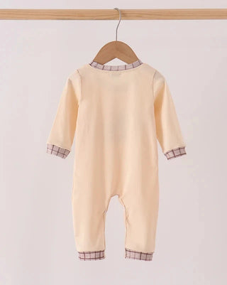 A pale peach baby jumpsuit hanging on a wooden hanger from a light wood rod, featuring subtle plaid trim at the collar, cuffs, and hems.