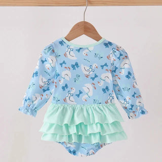 A baby long-sleeve romper hanging on a wooden hanger against a light beige wall. The top is light blue with a pattern of white swans and blue butterflies, and the bottom features a mint green ruffled tiered waistband.