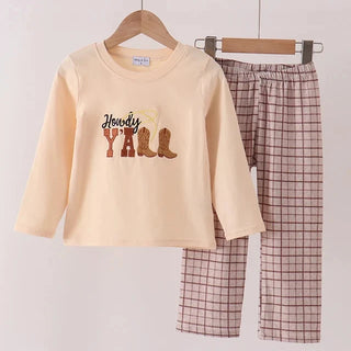 A two-piece children's outfit hanging on wooden hangers against a plain light background. The set includes a long-sleeve cream-colored top with a 'Holidays Y'all' graphic featuring cowboy boots, and matching plaid pants in a muted pinkish-brown and beige pattern.