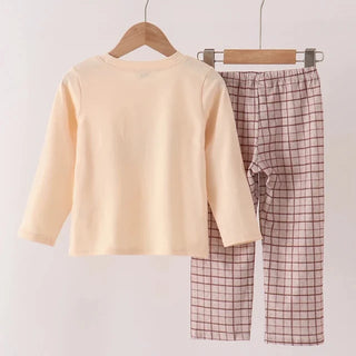 A children's two-piece pajama set hanging on wooden hangers from a wooden rod. The set includes a pale peach long-sleeve top and matching pink and white checkered pants.