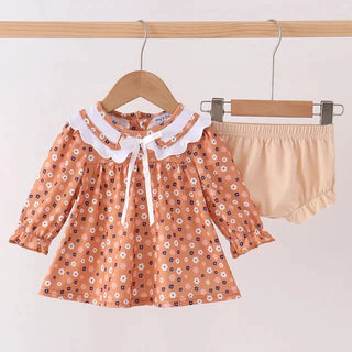 A toddler girl's two-piece clothing set hanging on wooden hangers against a plain white wall. The set includes a long-sleeve blouse in a peach-orange color with a white floral pattern, a white ruffled Peter Pan collar, and a white bow at the neck, paired with beige ruffled bloomers.