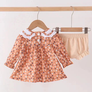A toddler girl's two-piece outfit set hanging on wooden hangers against a light beige wall. The set includes a long-sleeve blouse in a peach-orange color with a white and blue floral pattern and a white ruffled collar, paired with beige bloomers.