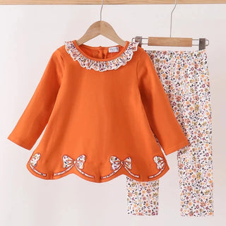 A girl's two-piece clothing set hanging on wooden hangers against a plain white wall. The set features a bright orange long-sleeve top with a white floral ruffled collar and bow details along the hem, paired with matching white floral print pants.