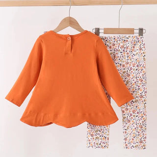 A toddler girl's two-piece outfit hanging on wooden hangers against a plain white wall. The set includes a bright orange long-sleeve peplum-style top with a single button at the back neckline, and matching floral print pants with a white base and small multicolor flowers.
