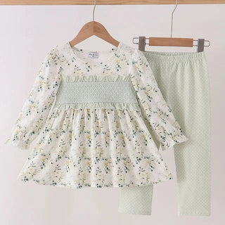 A girl's two-piece clothing set hanging on wooden hangers against a plain light background. The set includes a long-sleeve smocked blouse in an off-white fabric with a delicate green and yellow floral print, and matching light sage green polka-dot leggings.