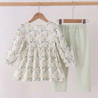 A two-piece children's outfit hanging on wooden hangers against a plain light background. The set includes a long-sleeve white blouse with a delicate green and yellow floral print and matching green leaves, paired with light sage green polka-dot pants.