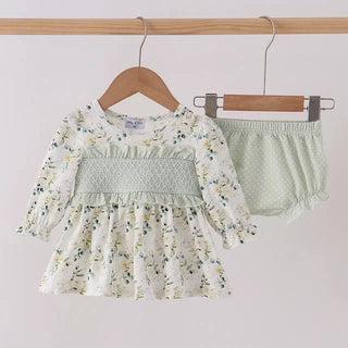 A baby girl's two-piece outfit hanging on wooden hangers against a plain light background. The set includes a long-sleeve smocked dress in an off-white floral print with green and yellow details, and matching light sage green ruffled bloomers.