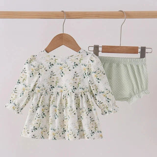 A two-piece baby clothing set hanging on wooden hangers from a wooden rod. The set includes a long-sleeve white top with a delicate green and yellow floral print, and a pair of light sage green smocked bloomers with a scalloped edge.