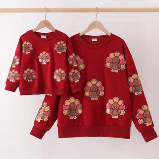 A matching mother-and-child red sweatshirt set hanging on wooden hangers against a plain white wall. The larger long-sleeve sweatshirt and the smaller cropped sweatshirt both feature an all-over pattern of cartoon turkeys in red, yellow, orange, and brown.