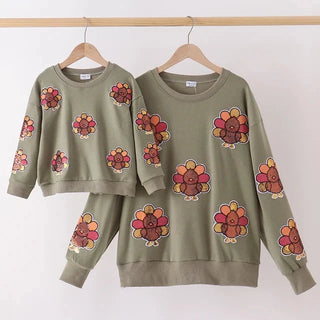 A matching mother-and-child sweatshirt set in sage green, featuring a colorful turkey print. The set includes a cropped long-sleeve top for the child and a long-sleeve sweatshirt for the mother, both hanging on wooden hangers from a light wood rack against a plain white wall.