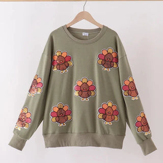 An olive green crewneck sweatshirt with a repeating pattern of cartoon turkeys in orange, yellow, and red, hanging on a wooden hanger against a plain white wall.