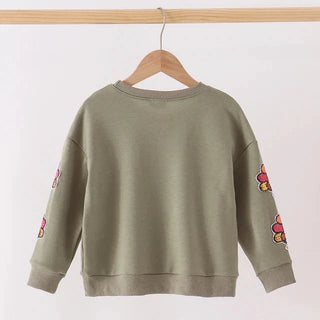 An olive green crewneck sweatshirt with colorful floral embroidery on both sleeves, hanging on a wooden hanger from a light wood rod against a plain white background.
